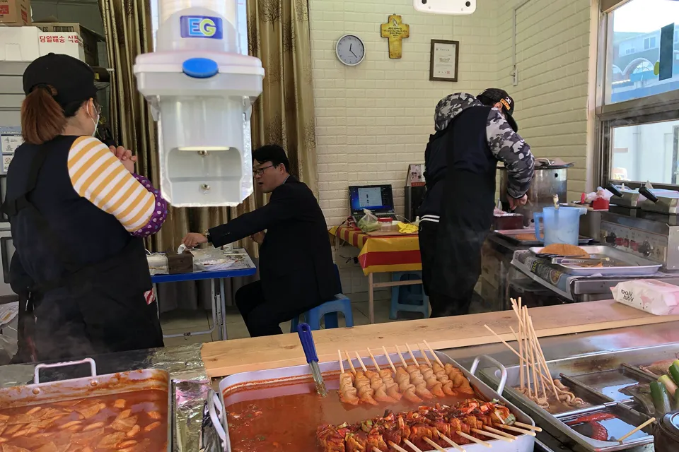 작은 분식집 카운터에 떡볶이와 어묵꼬치가 놓여 있고, 안쪽에 일하는 사람과 앉아 있는 손님이 보이는 모습